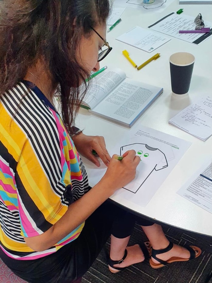 Looking over the shoulder of a course participant with long dark hair, as she sits at a white desk, drawing a green smiling face on a page with a green texta. 