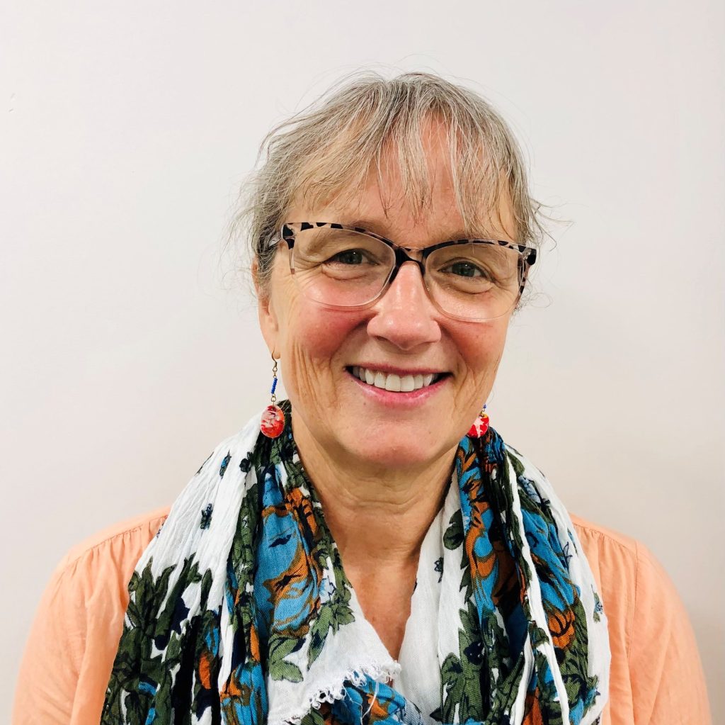 Mental Health Consultant Engel Prendergast smiles warmly at the camera. Engel wears tortoise shell glasses, red and gold earing, a white floral scarf and a peach shirt.