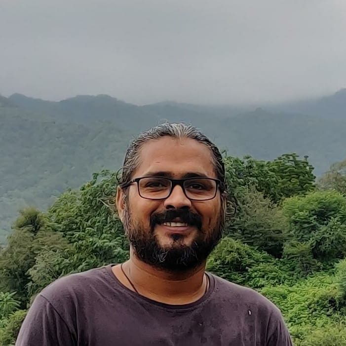 Mahaveer smiles warmly at the camera, with a foggy, green forest mountain range in the background. Mahaveer wears black glasses, a short, dark beard and a grey t-shirt.