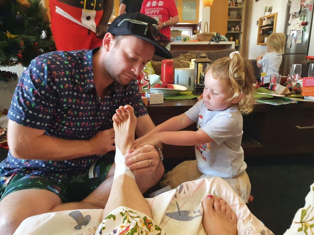 Little blonde girl lovingly helping her dad strap her grandmother's sore ankle.