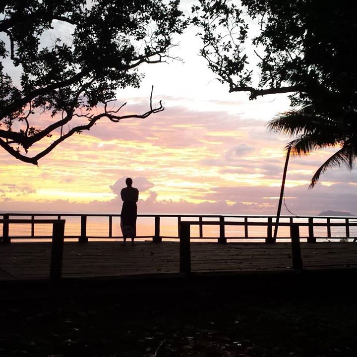 A woman stands on a foreshore - overlooking the sea at sunset, the sky gold and pink, filled with clouds.