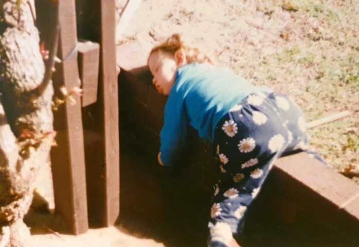 A toddler, wearing a mid-blue top and daisy-print blue leggings, lays asleep on a small red brick fence, exhausted from playing.