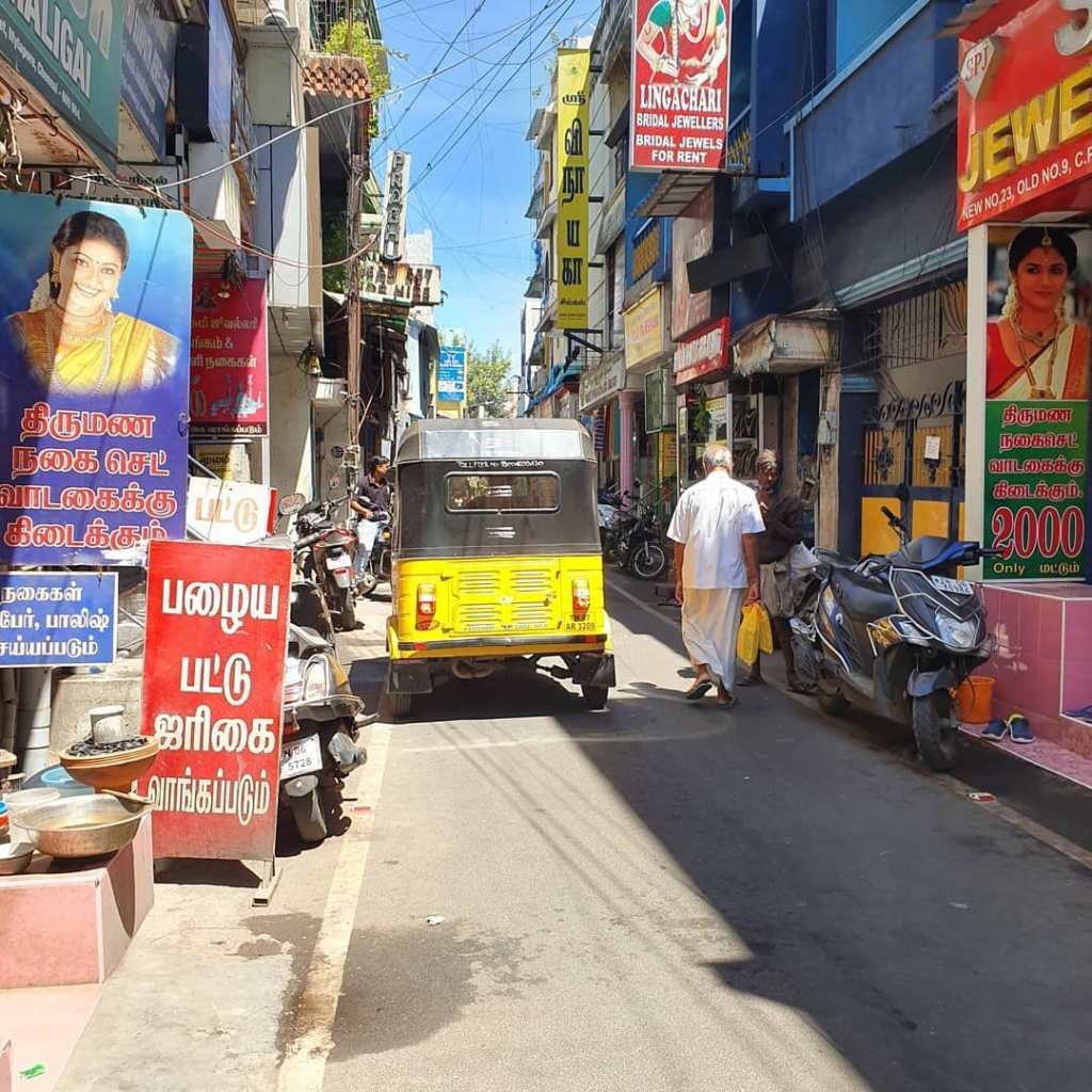 A yellow and black car drives down an Indian laneway. The laneway is filled with small shops, signs and motorbikes.