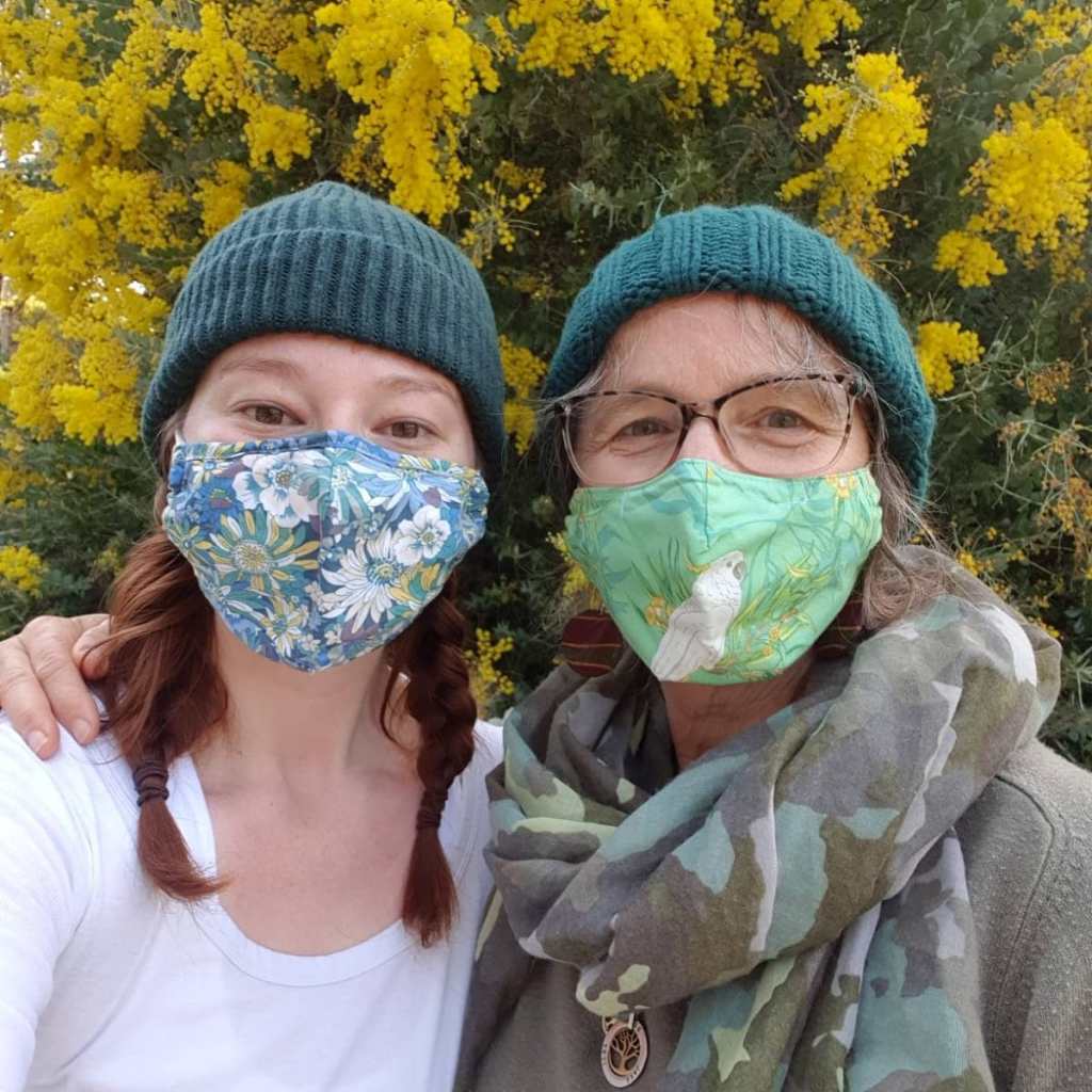 A mother a daughter, wearing green beanies and colourful masks, smile for the camera on a winter's day.