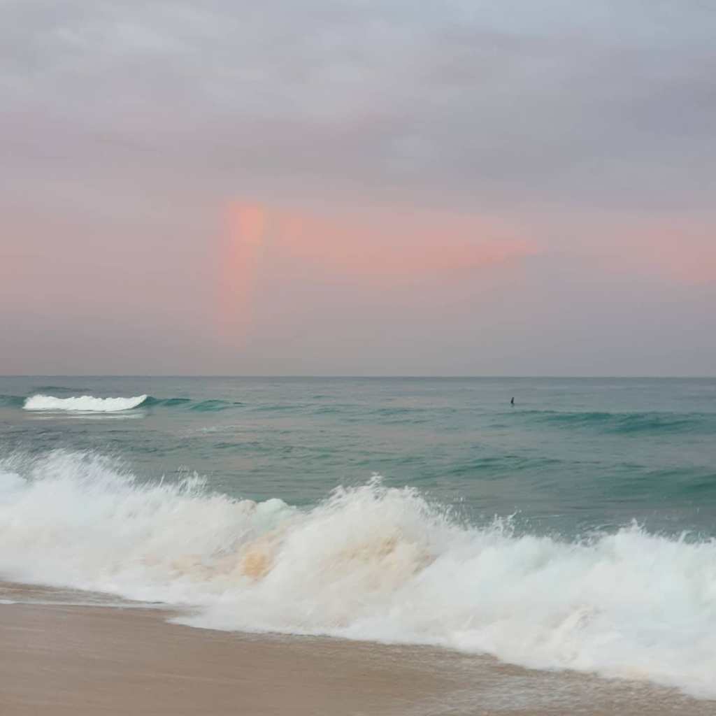 Rough waves break on a Perth beach at dusk, while a lone swimmer stands in the water beyond the waves.
