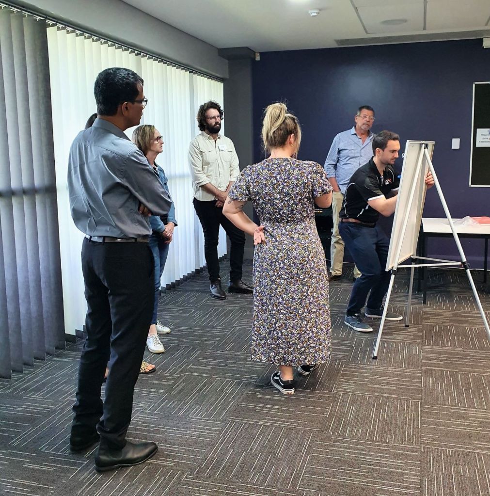 A group of seven (male and female) Mental Health First Aid course participants stand around a whiteboard on an easel. One man writes on the board while the others workshop ideas.