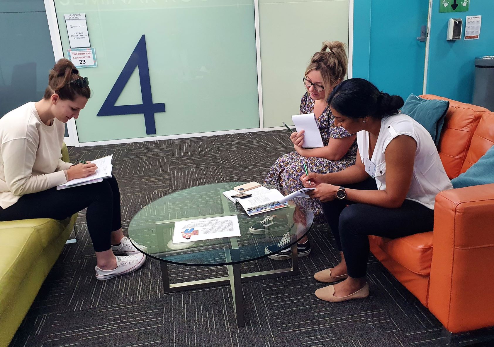 Three mental health course participants sit on a green and an orange couch, one leaning on a glass coffee table, workshopping course materials. 