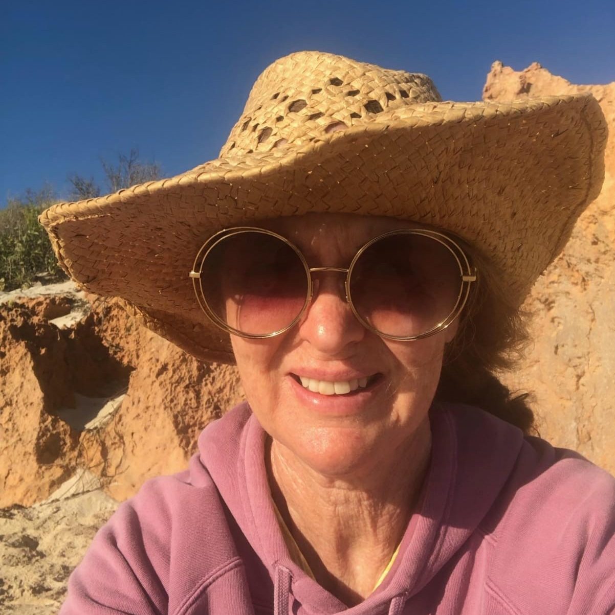 Cathy is at the beach - smiling at the camera, wearing a big straw hat, oversized, gold sunglasses, and a violet hooded jumper.