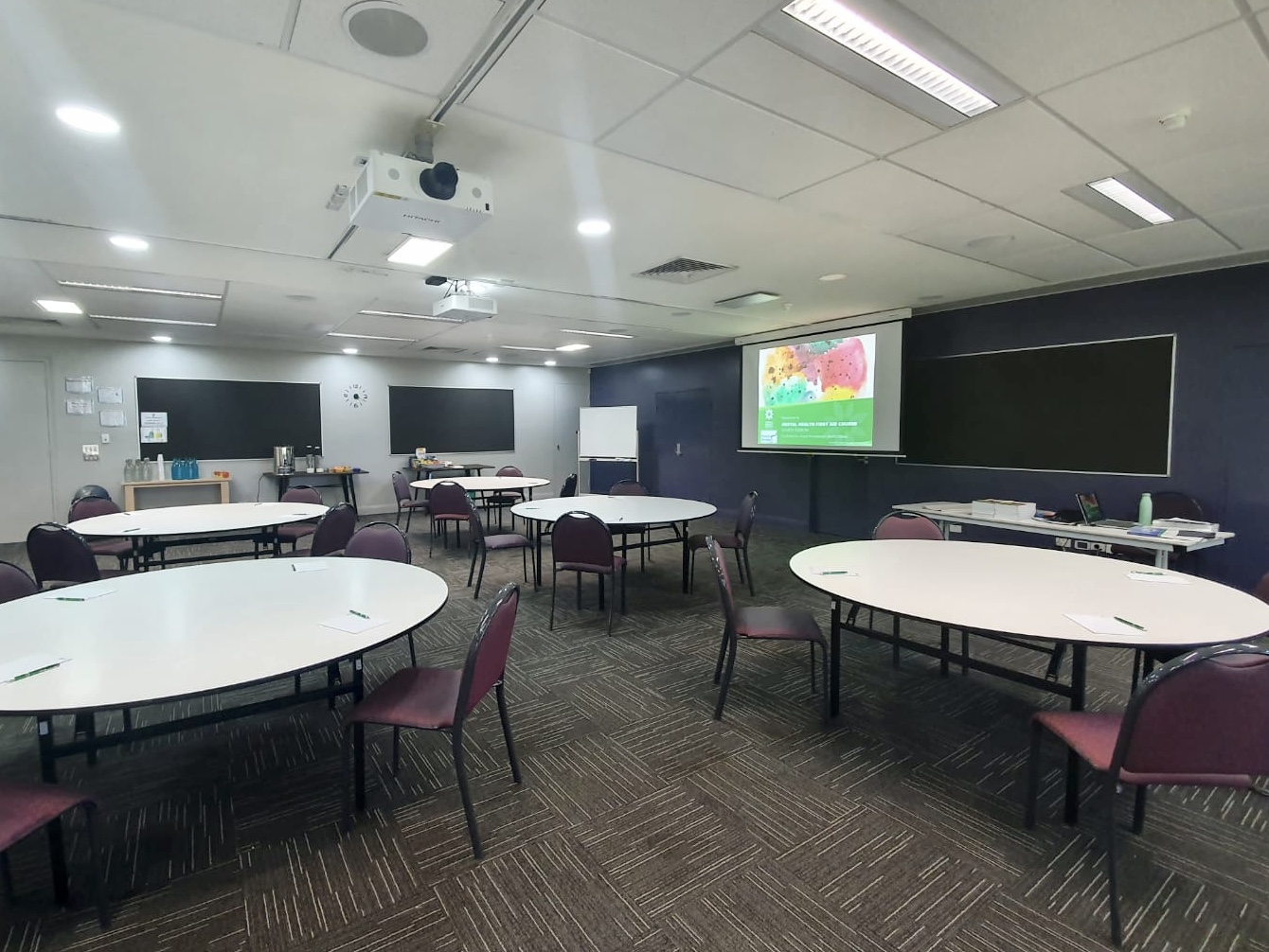 A corporate training room, with five round tables and purple chairs, pens and notepads on each desk, with a projectors screen at the front of the room featuring the cover page of the Mental Health First Aid training presentation. 