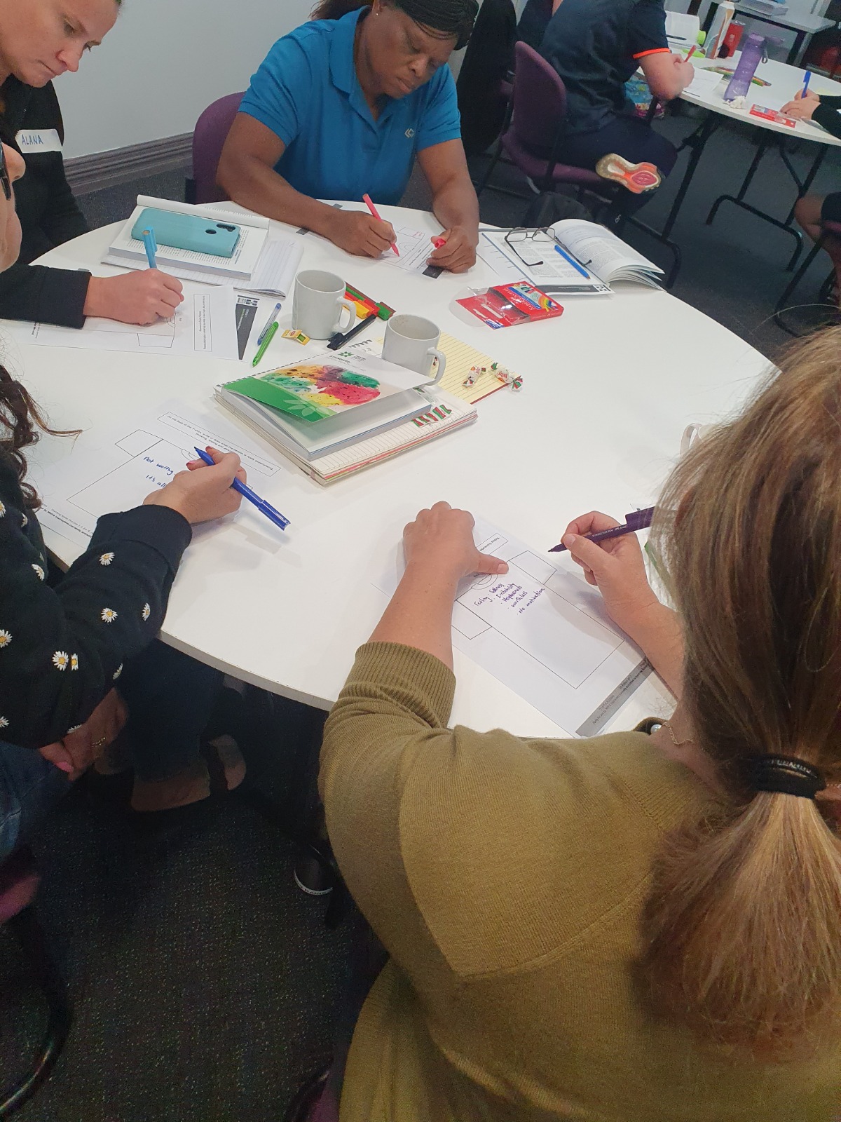Four people sit around a white table with pens in hand, writing as part of a Mental Health First Aid training activity. 