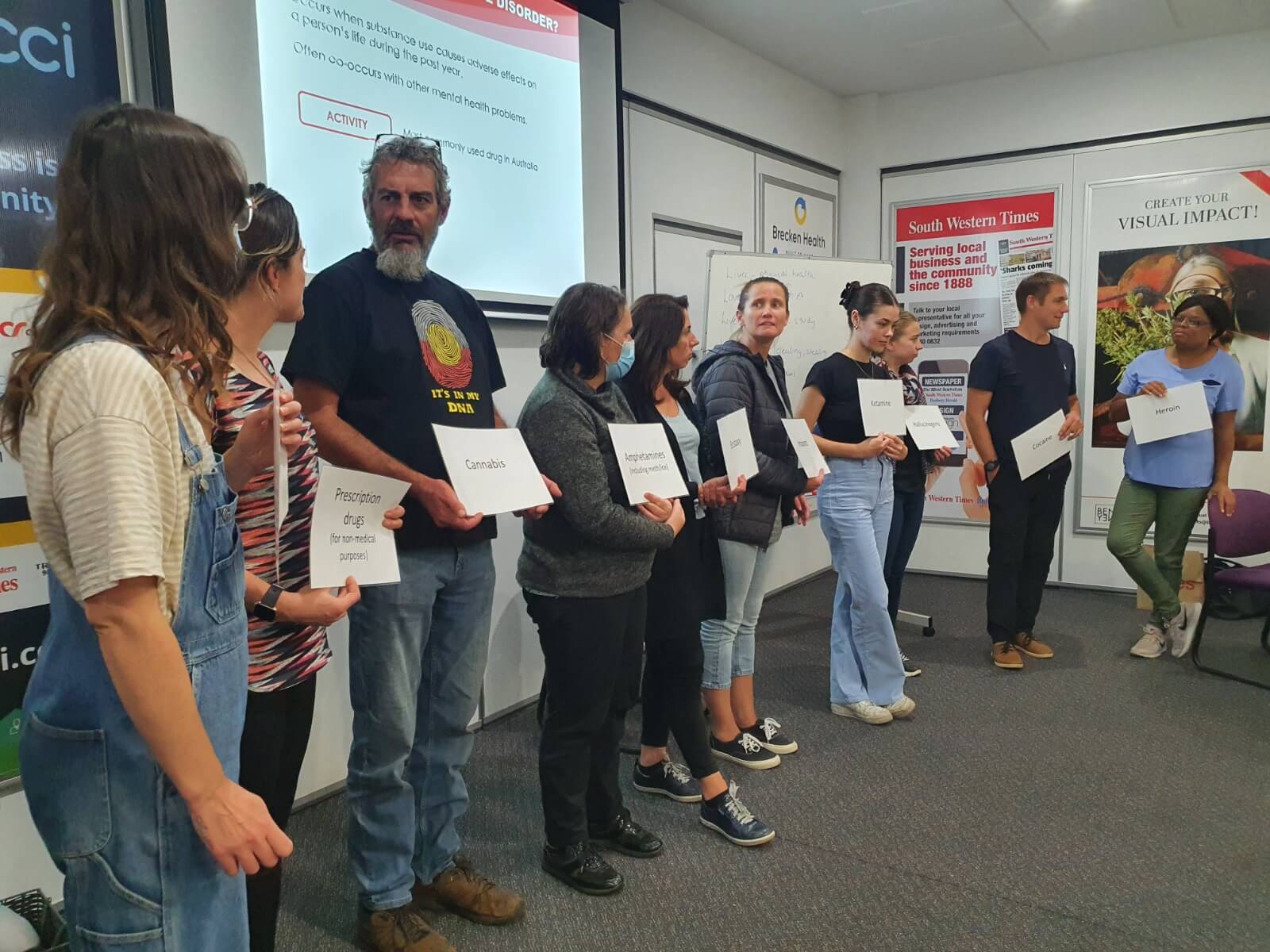 A group of ten men and women stand together at the front of a room, chatting and holding A4 white pages, each featuring the names of types of drugs as part of a a Mental Health First Aid training activity.