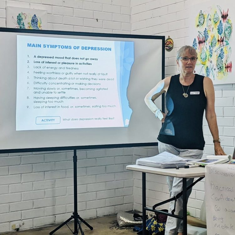 Mental Strides' Mental Health Consultant, Engel Prendergast stands in front of a projector screen, wearing a black top and glasses to deliver mental health training. 