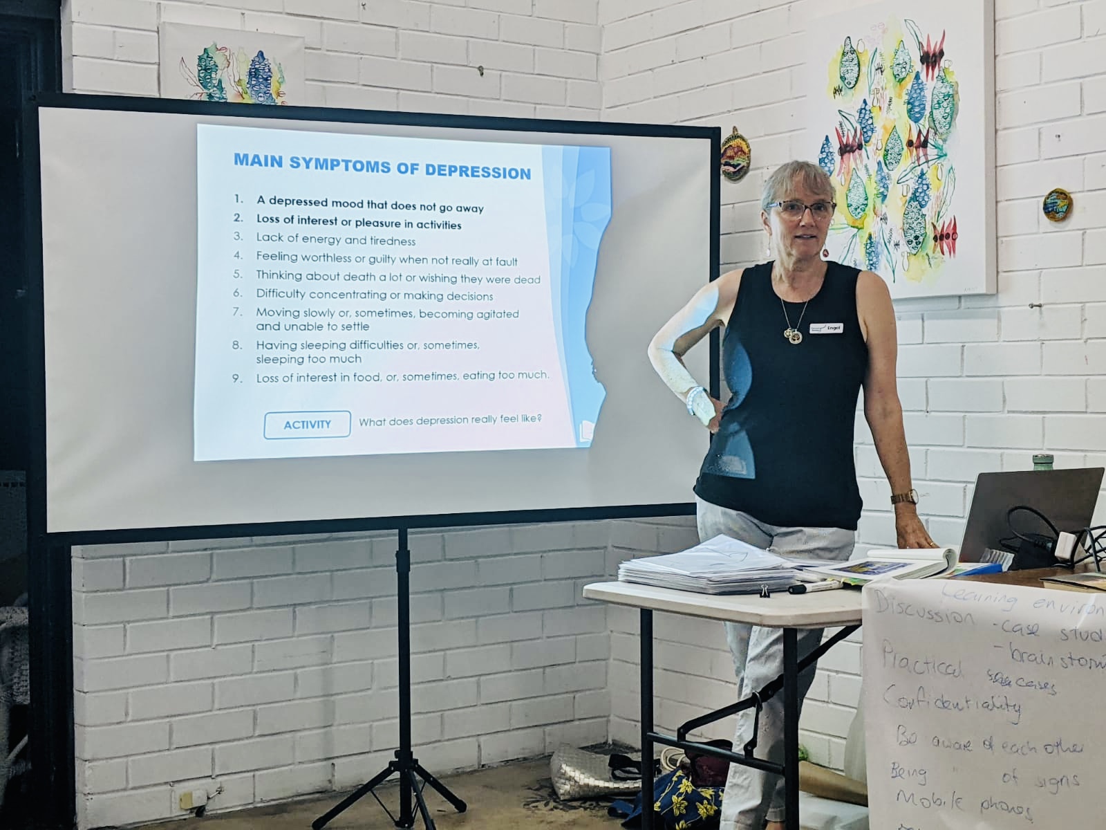 Mental Strides' Mental Health Consultant, Engel Prendergast smiles warmly at the camera. She has her hair back, and wears tortoise shell glasses, a black sleeveless top and grey pants. Engel is standing at the front of a room, delivering Mental Health First Aid training. A projector behind her features a tile titled, "Main Symptoms of Depression".