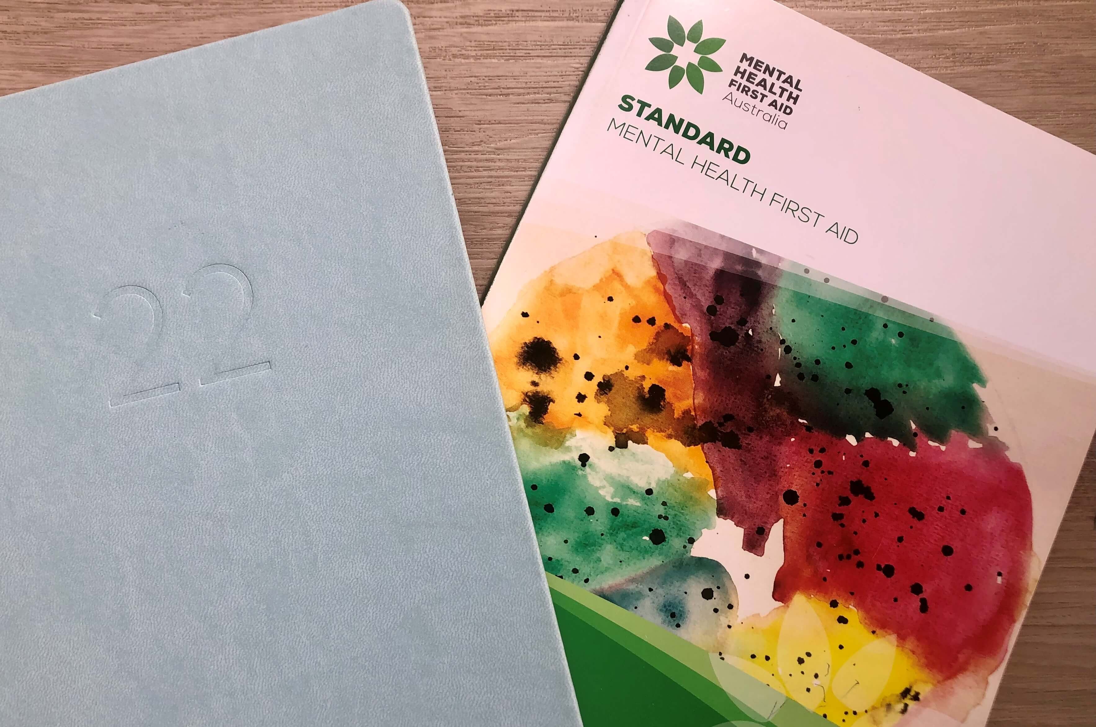 A light blue 2022 diary and the Standard Mental Health First Aid course book, sitting on a light oak table. 