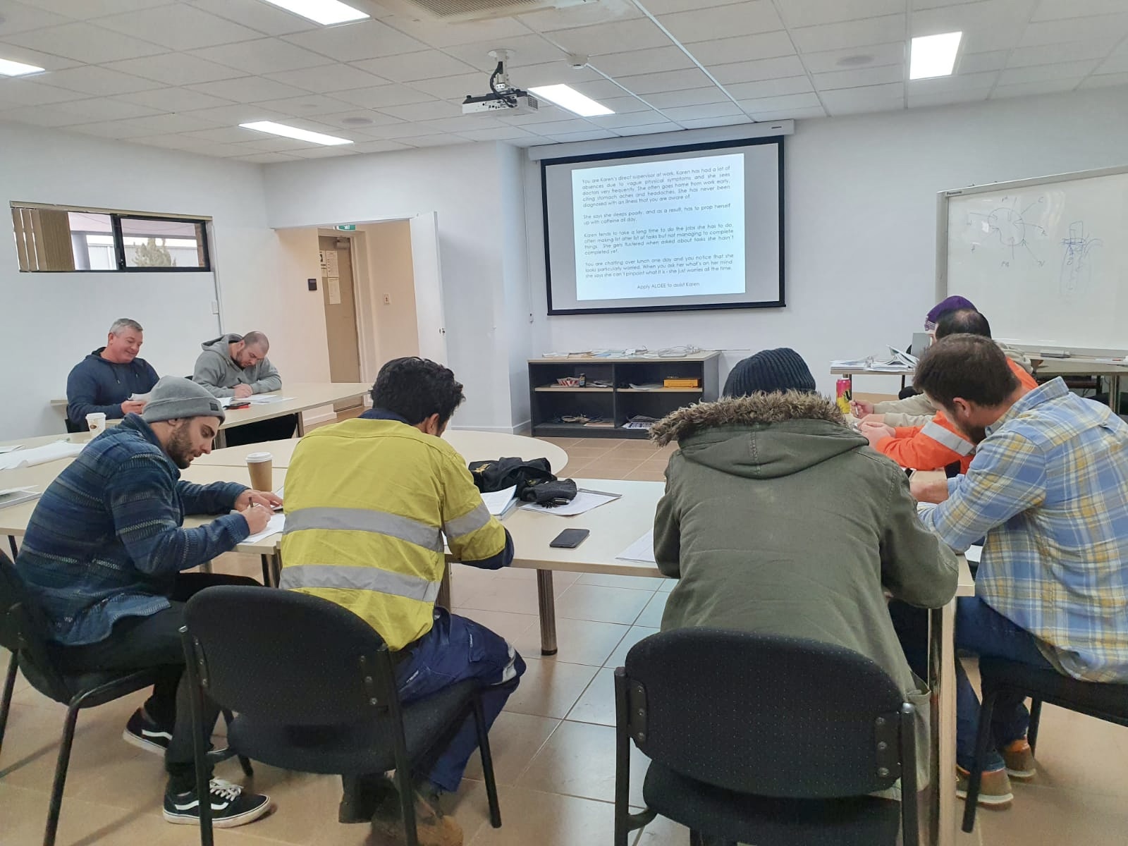 Eight men sit around a u-shaped group of tables, writing part of a Mental Health First Aid training activity. A projector screen is at the front of the room with course information on display. 
