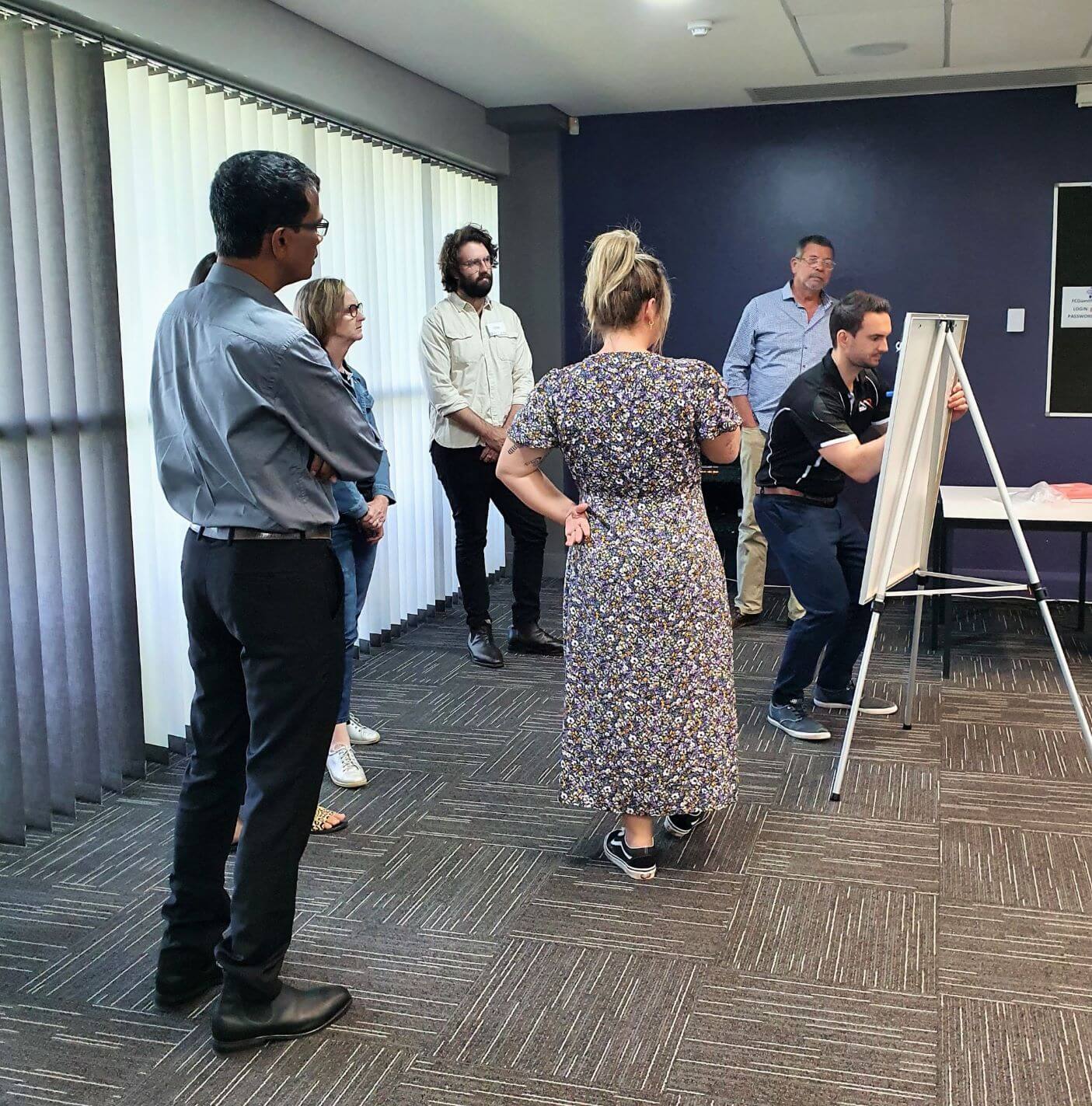 Seven Mental Health First Aid course participants stand around a whiteboard on an easel. One man writes on the board while the others look on, workshopping ideas as part of a course activity.