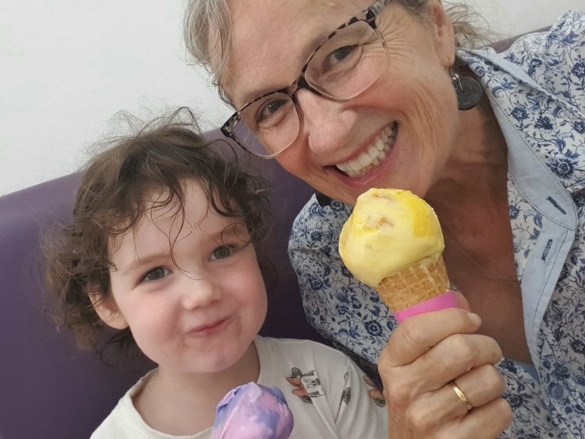 Engel wears a white and blue button-up shirt and tortoise shell glasses, whilst eating a mango flavoured scoop of ice-cream in a cone. A brown haired toddler smiles alongside her whilst holding a purple, bubble-gum flavoured ice-cream in a cone. 