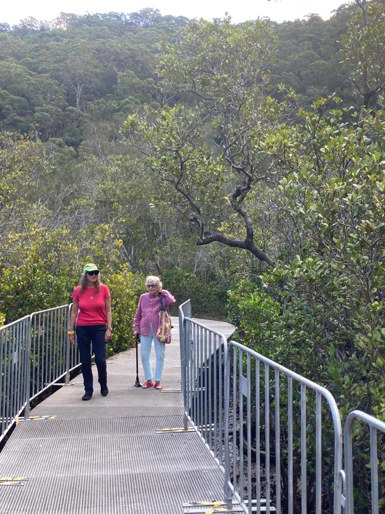 Engel wears a lime green cap. hot pink t-shirt, black pants and black sneakers alongside her mother, who wears a dusty pink shirt and light blue pants, and carries a bright coloured bag. They walk along a metal boardwalk with side railing in the Australian bush. 