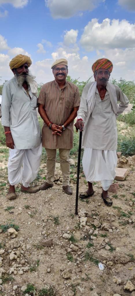 An Indian man in a brown and orange striped shirt and cream hat stands in between two Indian elders, one supports himself with a dark wood walking stick. Both elders wear a pagri on their heads, and traditional, light-coloured long shirts and pants. 