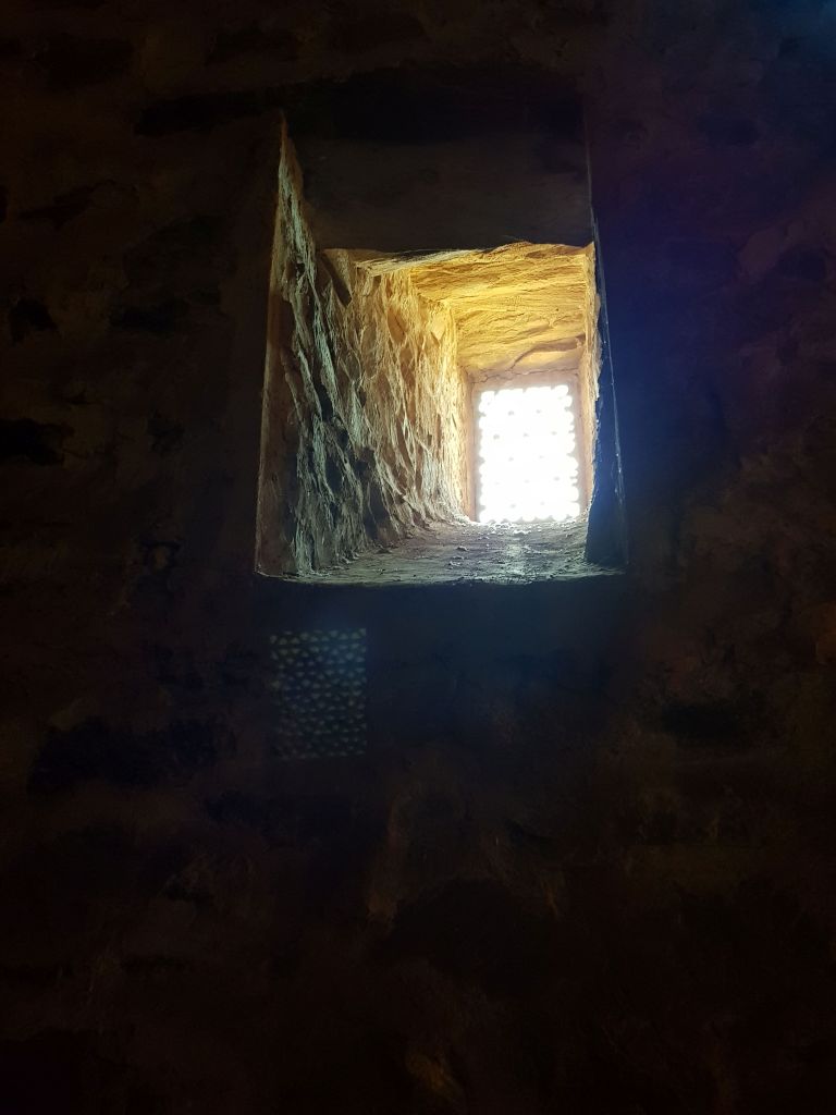 Looking up out of small, concrete window from a very dark, thick limestone room. The bright light shines through in a direct, square shape and you can see the colour of the limestone in the window surrounds.