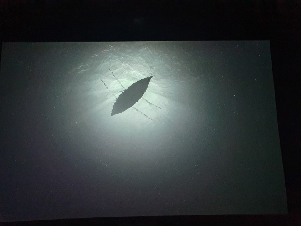 Looking up from under the dark sea towards a row boat with six oars, lit by the moonlight above.
