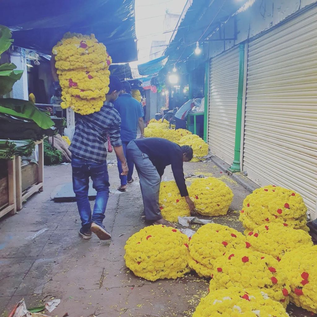 Two men in India lift and carry large arrangements of yellow flowers at a market. 