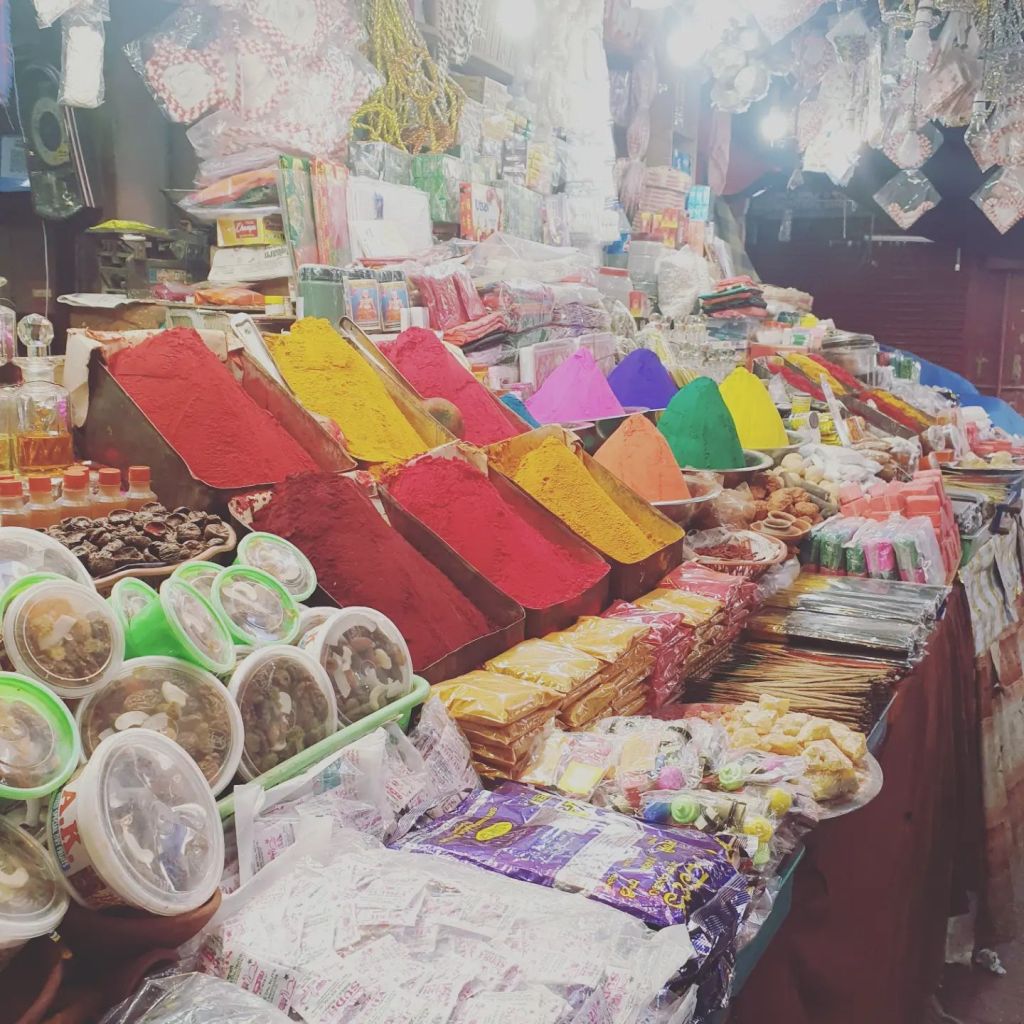 Indian market stall featuring food and bright-coloured spices in shades of red, mustard, yellow, pink, purple, green.