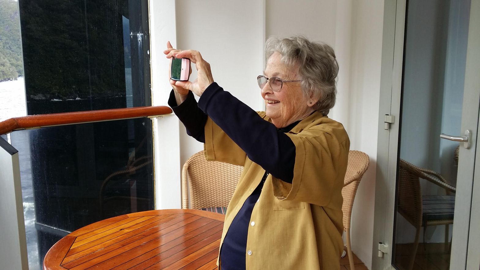 A lively and spritely mature lady holding a mobile phone in front of her to take a photo in the distance while standing on a balcony.