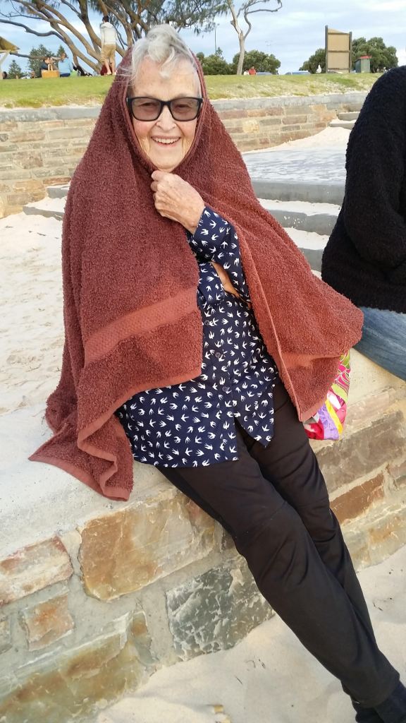 A spritely woman in her 90s wears tinted glasses and a towel around her head and body to shield herself from the weather.