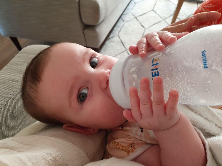 A brown-haired baby lays back, drinking a bottle of milk, looking at the camera while laying on their grandmother's knee.